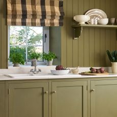 Green farmhouse kitchen with green painted kitchen cabinets and slatted wall panelling. Green gingham blinds hang over the window.