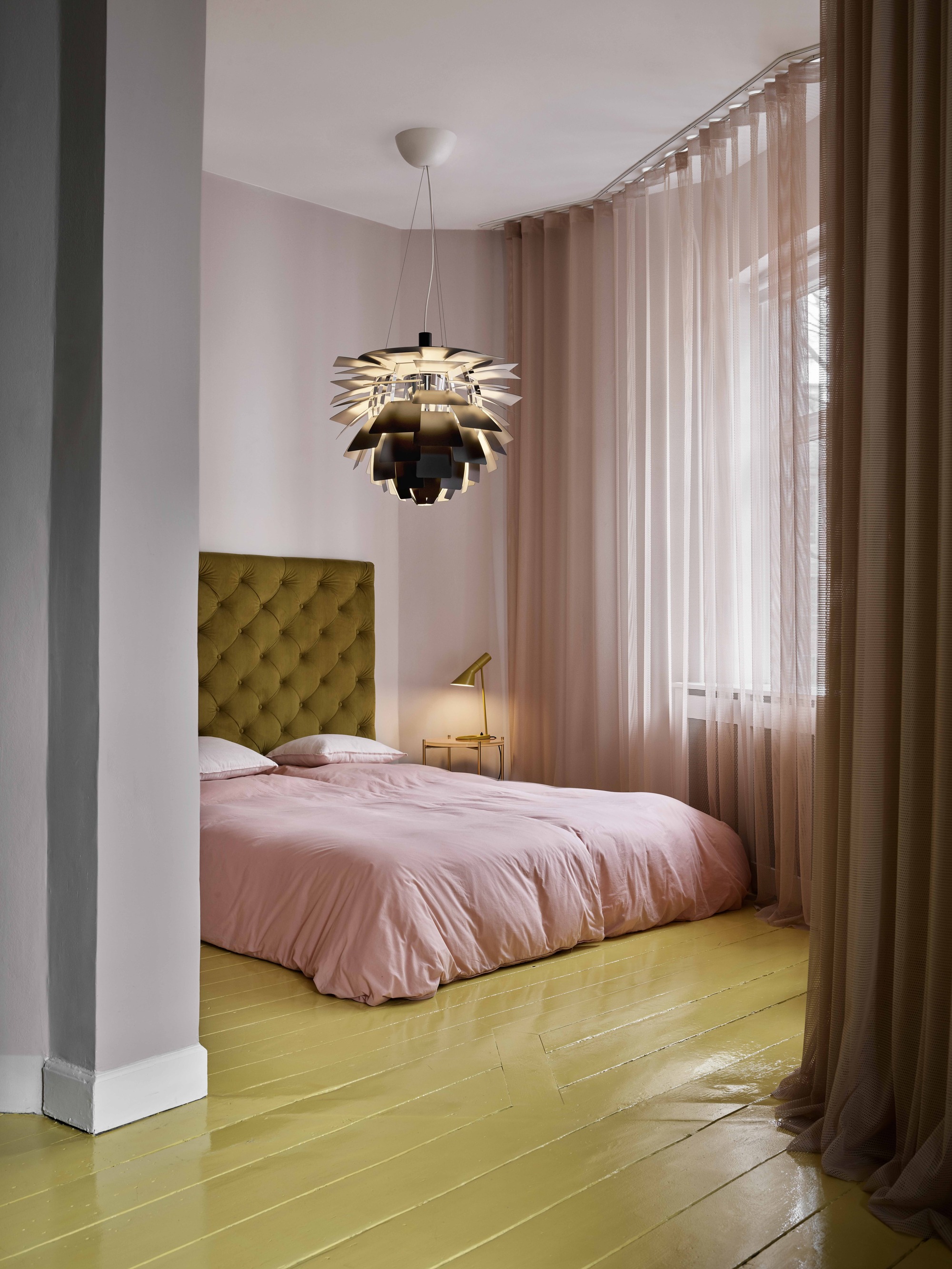 a colorful bedroom with bright yellow floors, pink bedding, pink walls and sheers, a louis poulsen pendant light and tufted upholstered mustard yellow headboard