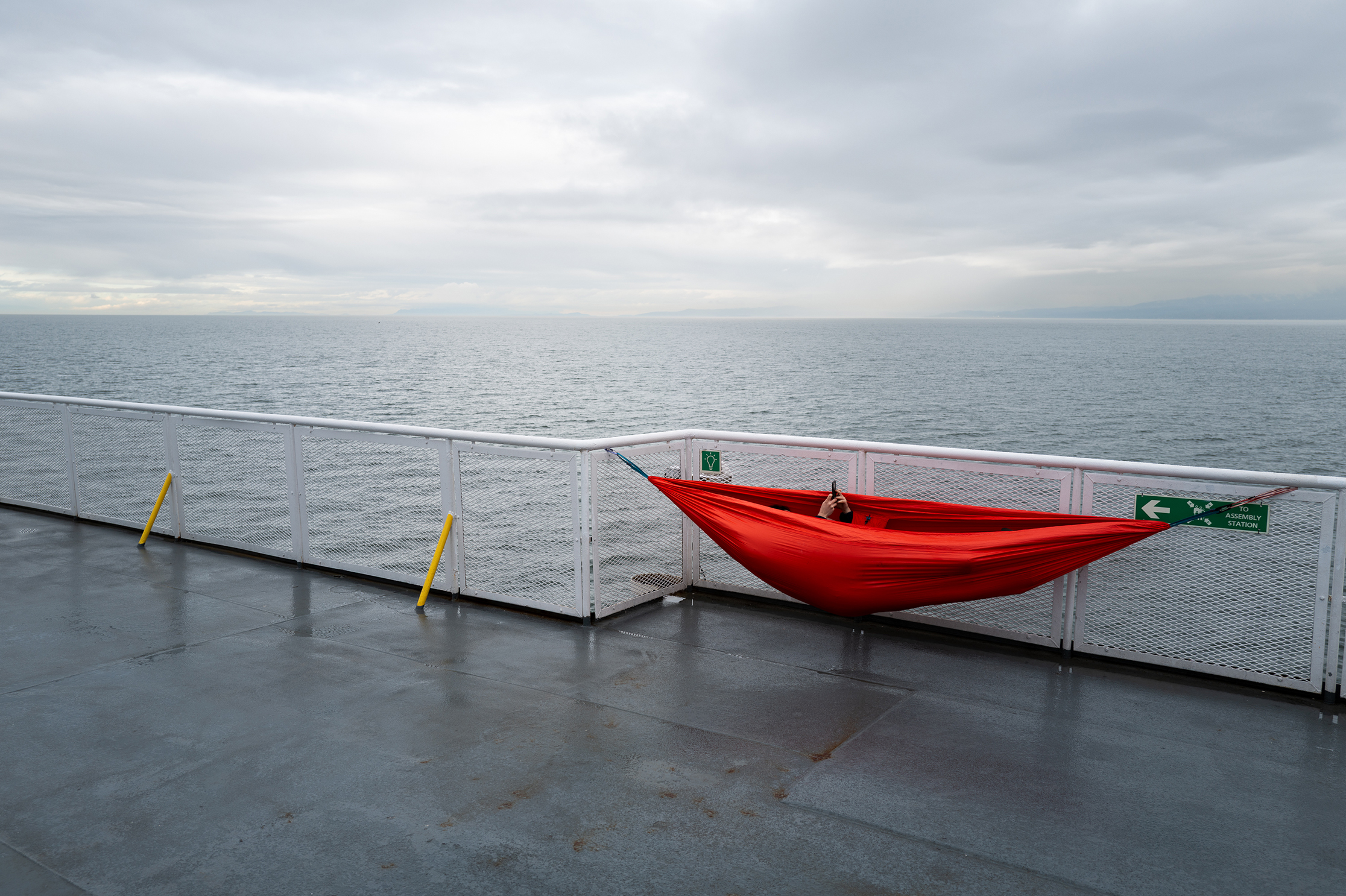 Red hammock stretched across a ferry deck railing overlooking a gray, choppy sea under an overcast sky