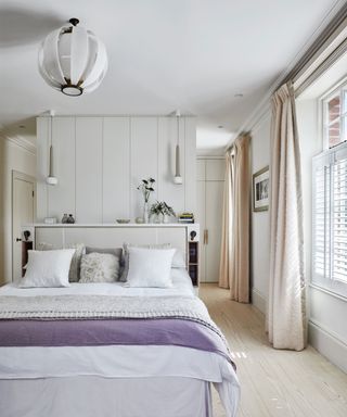 A light and bright bedroom with a white bed with a purple throw under a white, layered ceiling light