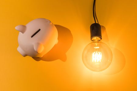 Piggy bank next to lit light bulb on pink background