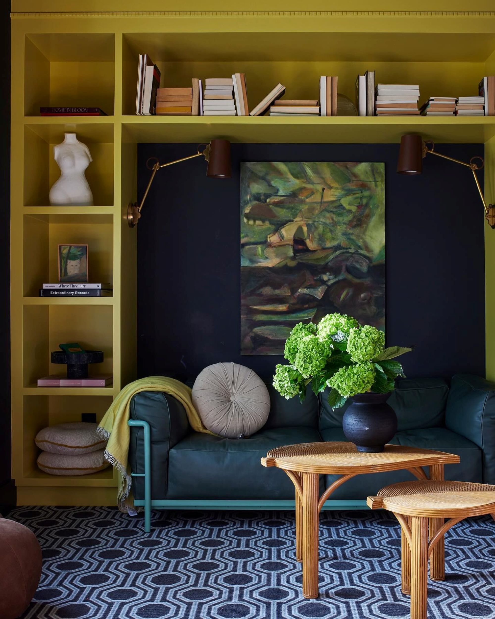 a living room with a yellow-green built-in bookcase filled with miscellaneous books and objects, a blue accent wall, a blue couch with a round gray pillow, two coffee tables and one holding a plant, and a blue geometric rug