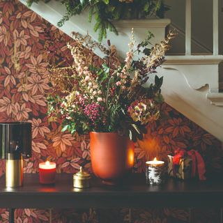 A hallway console table set against a leafy-print wallpaper with candles and a vase of flowers on top