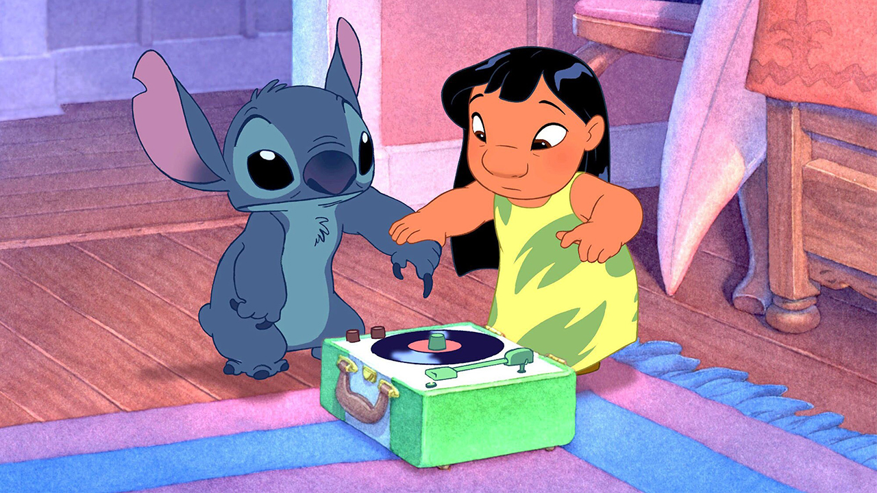 Still from the animated movie Lilo & Stitch (2002). A young Hawaiian girl is holding the hand of a blue, koala-like alien, hovering it's claw over a record player.