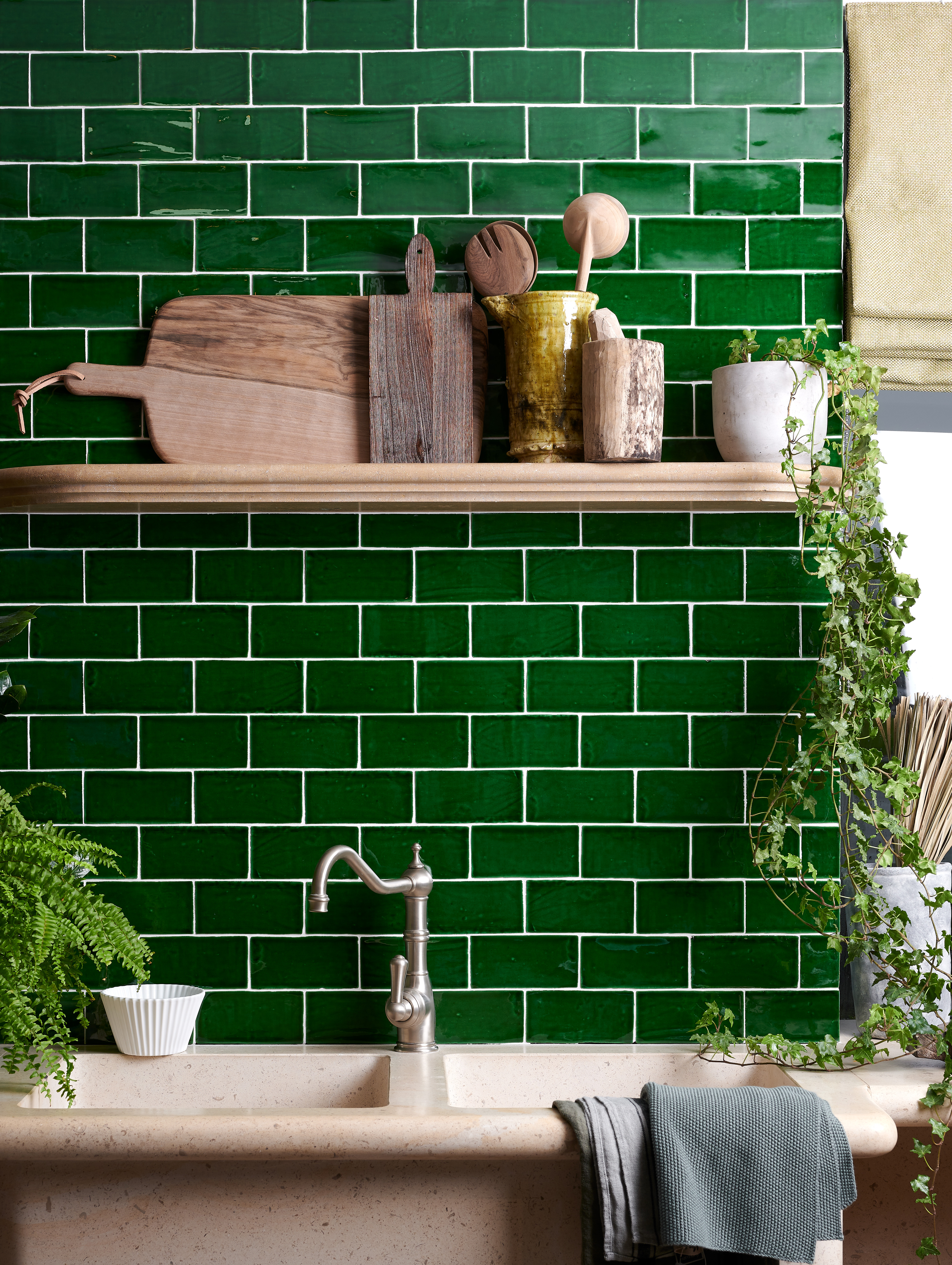 Green kitchen tiles behind a sink with wooden shelf.