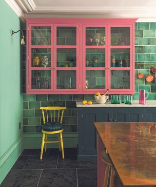 An example of small kitchen storage ideas showing pink, glass-fronted cabinets in front of a green-tiled wall and above a yellow chair