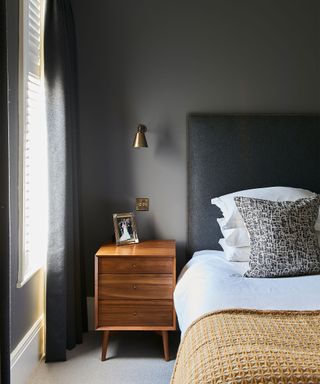 An example of bedroom wall lighting ideas showing a bed with a large grey headboard next to a small metal wall lamp and wooden bedside table.