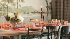 A dining area with a wood table set with rosey-pink glassware and placemats with a vase of flowers, raised serveware, and a trio of lit taper candles