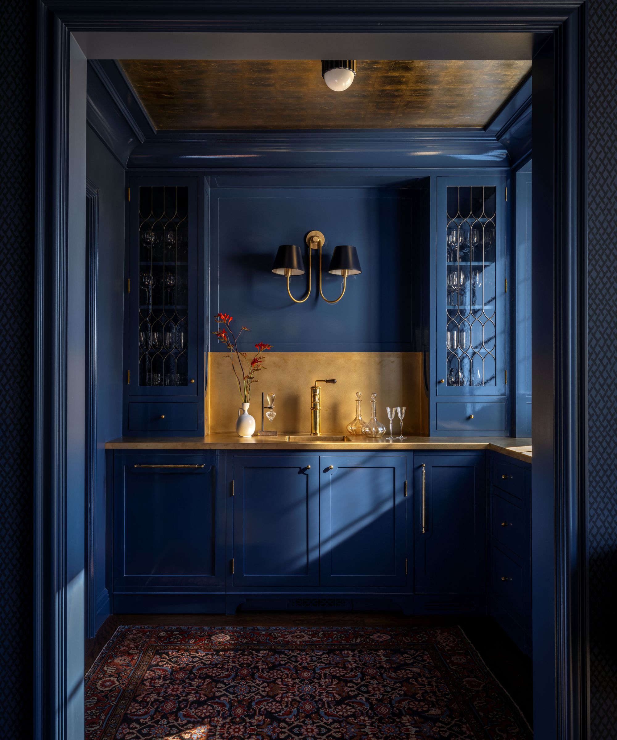 Bar pantry with dark blue painted cabinetry and walls, gold backsplash and countertops, glass cabinets with glassware, and a large blue sconce at the center with a red Persian rug on the floor