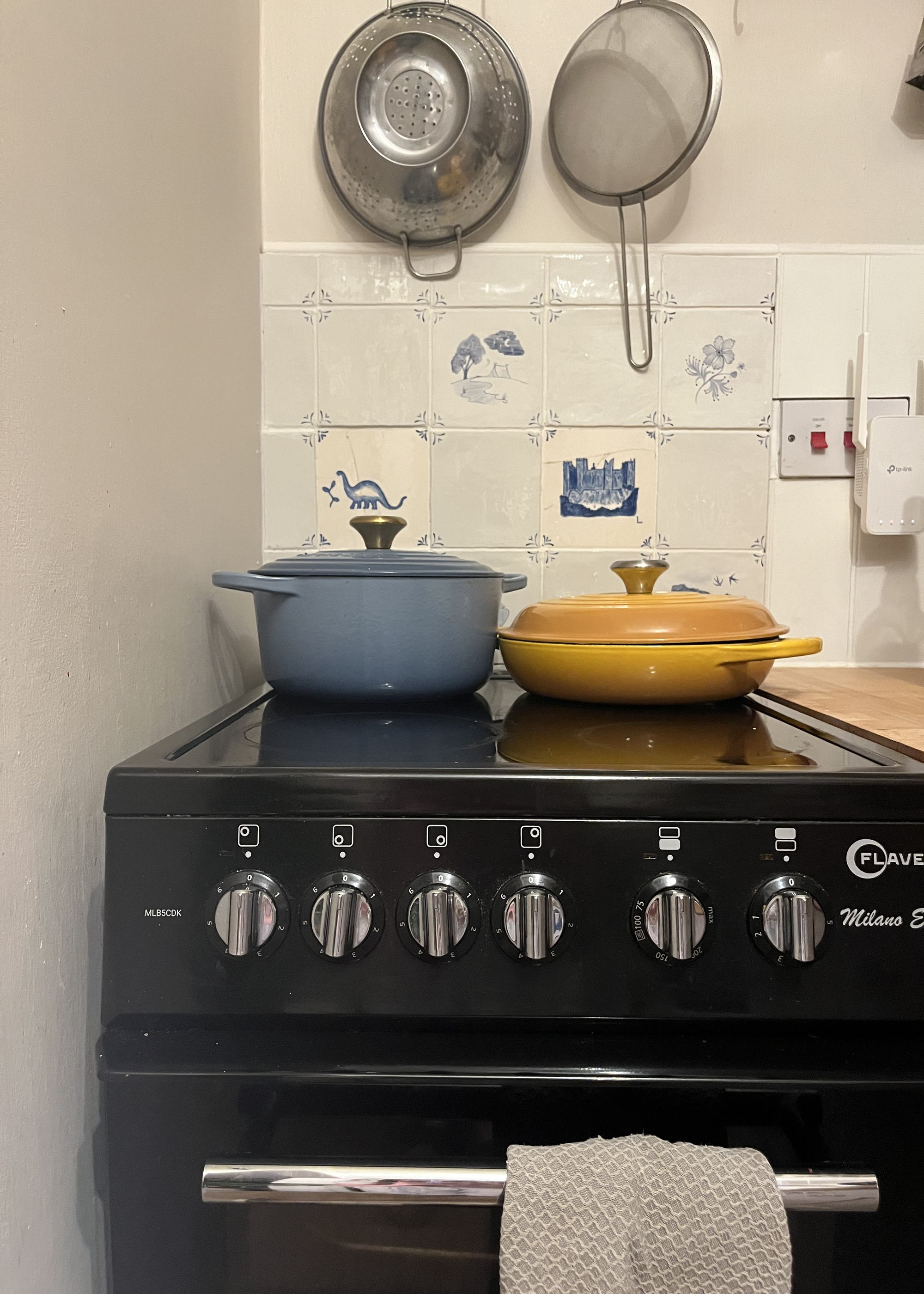 A kitchen with a black oven with two Le Creuset pans, one yellow, one blue, and a backsplash of Delft tiles
