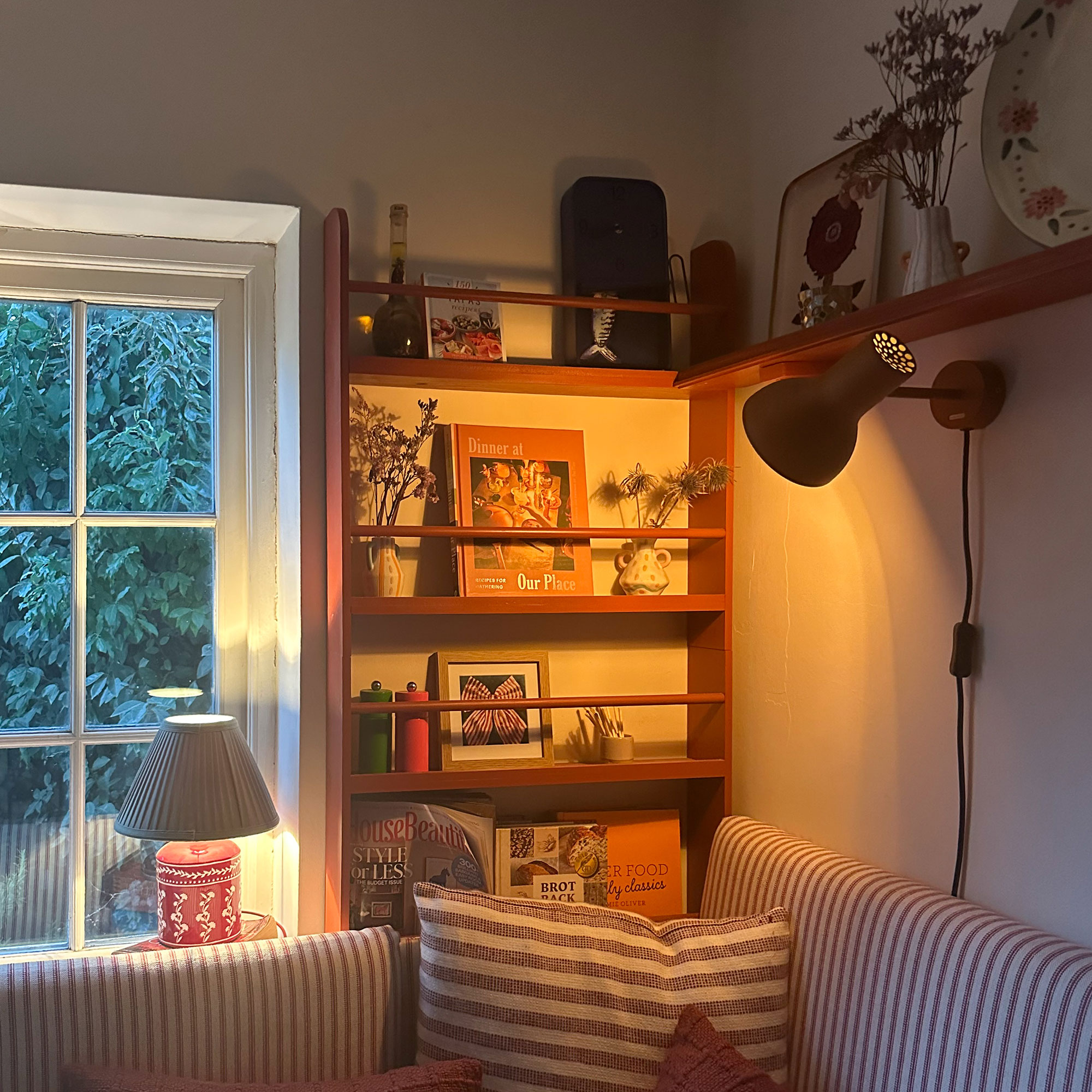 Corner of kitchen with red open shelving above red and white striped upholstered banquette, lit with wall and table lamps