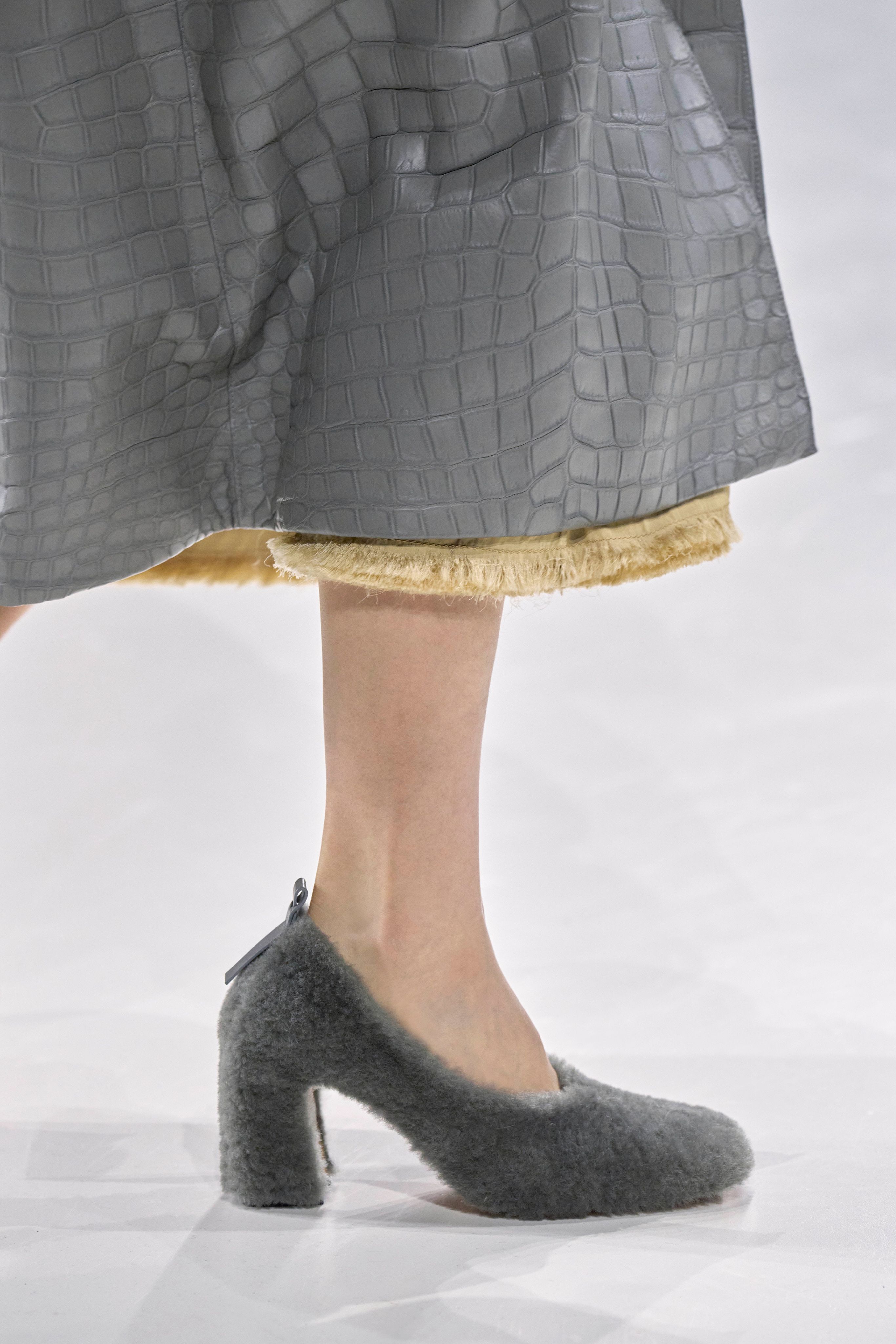 Fur accessory trend is shown in Bottega Veneta&#039;s spring summer 2025 collection in a close photo of a model&#039;s foot walking down the runway wearing shearling gray pumps with a yellow dress and a slate gray crocodile embossed leather coat