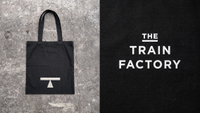 A black canvas tote bag printed with the white "T" logo laid on concrete, beside a close-up of "The Train Factory" wordmark embroidered in white on black fabric.