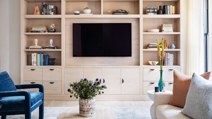 modern living room with built-in wall shelving, mounted tv, blue armchair opposite a white sofa with pillows, and a travertine coffee table with vase of flowers