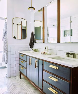 A white bathroom with three mirror cabinets, a navy blue and dark wood sink unit with white base, and white subway tiles on the wall and herringbone tiles on the floor