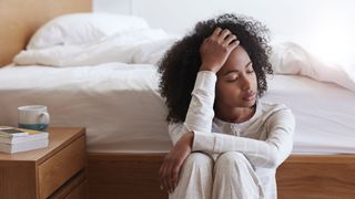 En kvinna sitter iklädd en vit pyjamas vid en sängkant med ena handen i pannan och ser trött ut.