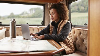 A woman sits at the table in a motor home with a map spread out before her and her laptop open.
