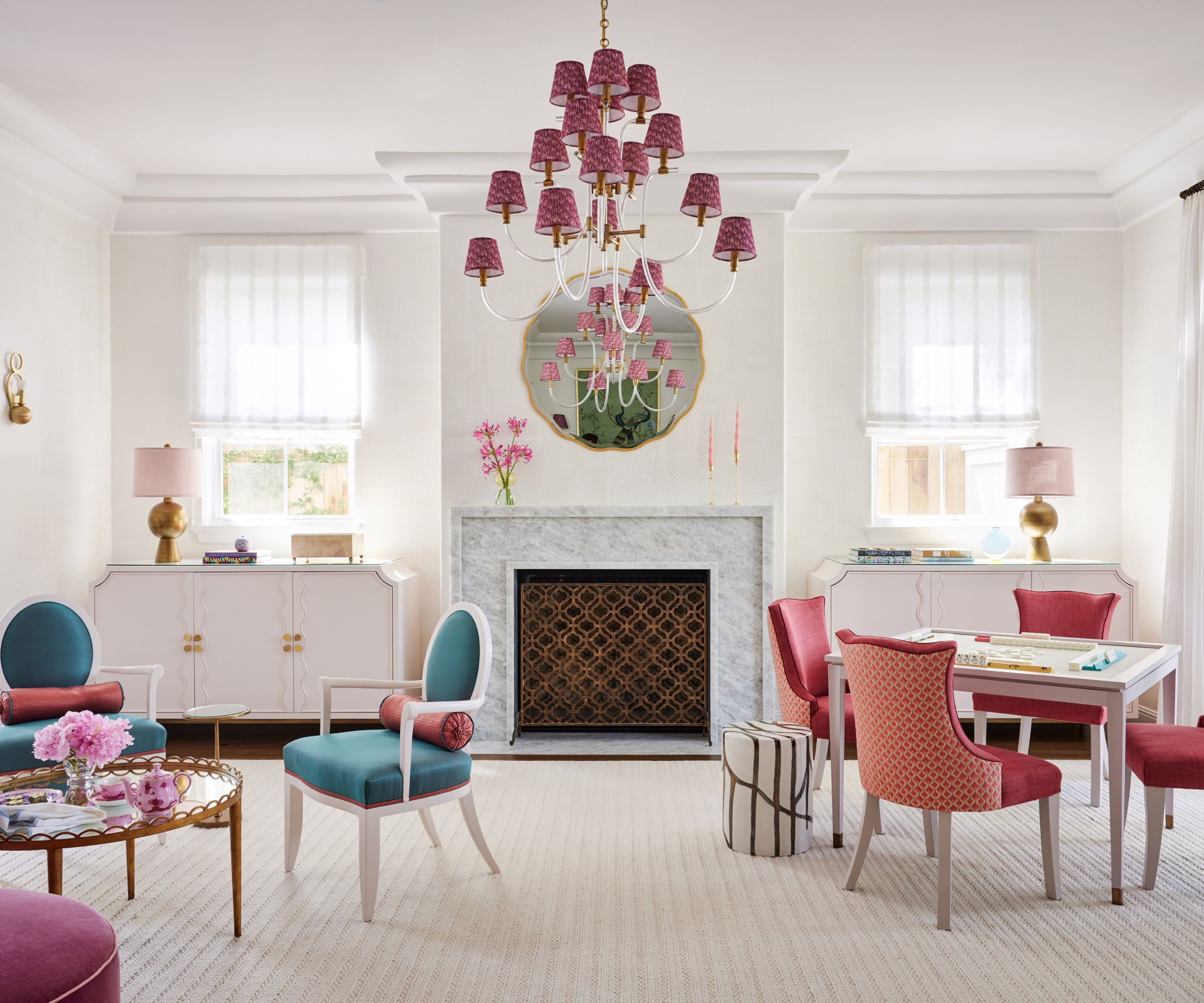 A white open plan room with two windows and a marble fireplace in between. There are white consoles within the alcoves, with matching old lamps. There are two seating areas, one with blue chairs and one with pink