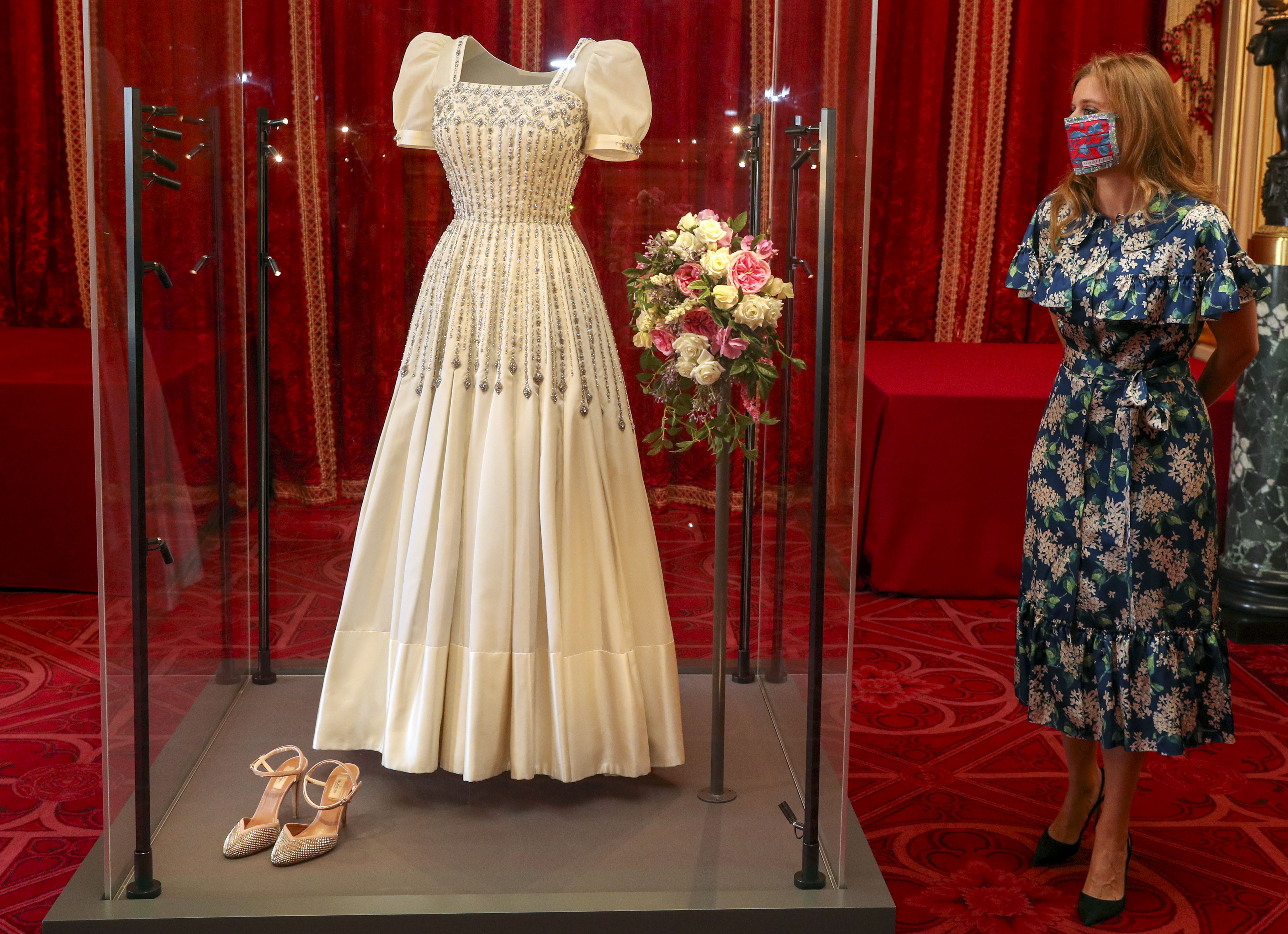 Princess Beatrice wearing a blue floral dress and face mask standing next to her wedding dress, which is in a display case