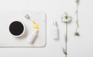 Sakhi organic ace oil bottle in all white placed next to a bowl with black seeds and a white flower.