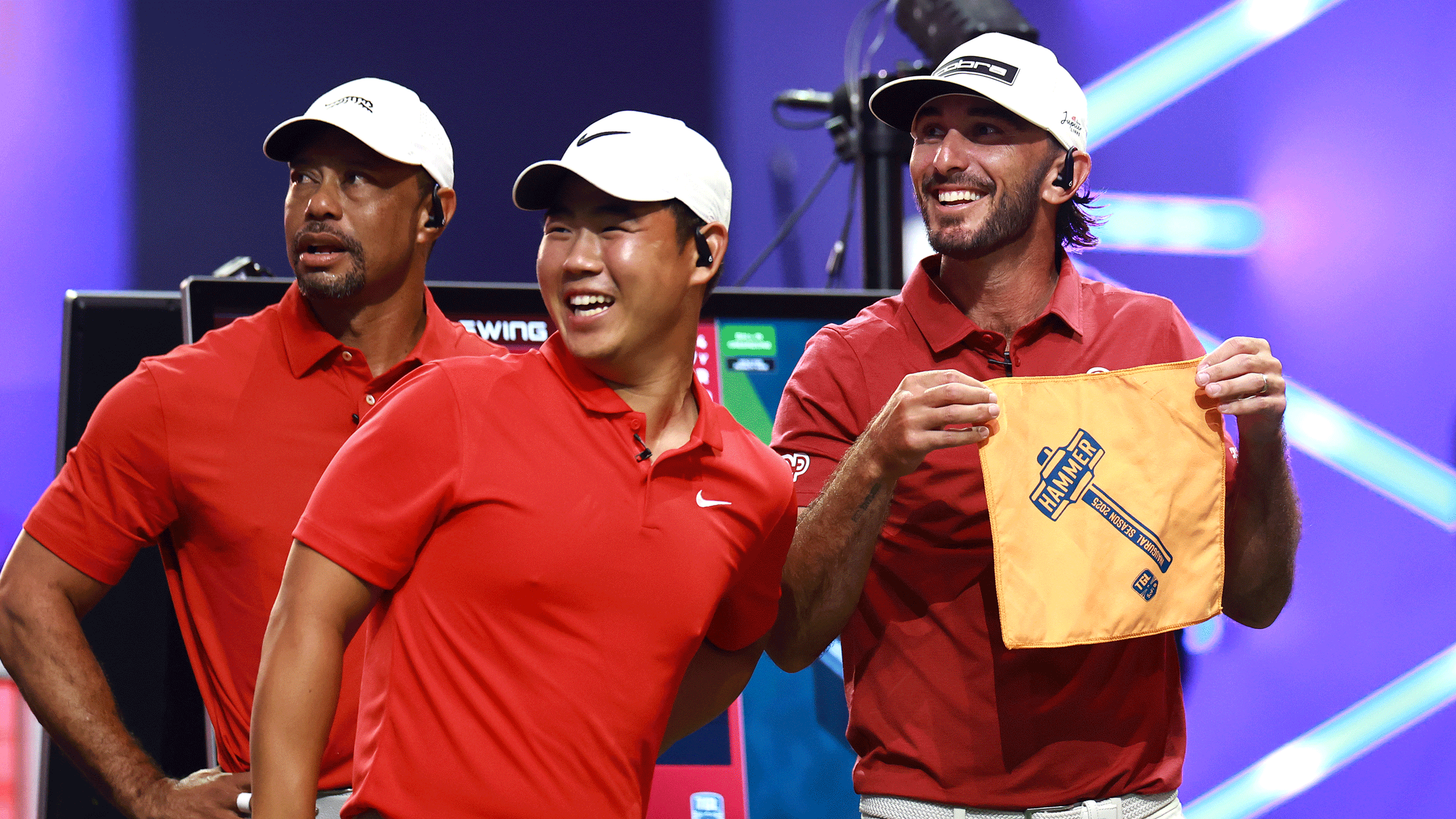 (L to R) Tiger Woods, Tom Kim and Max Homa look on while Homa holds up The Hammer flag in front of him