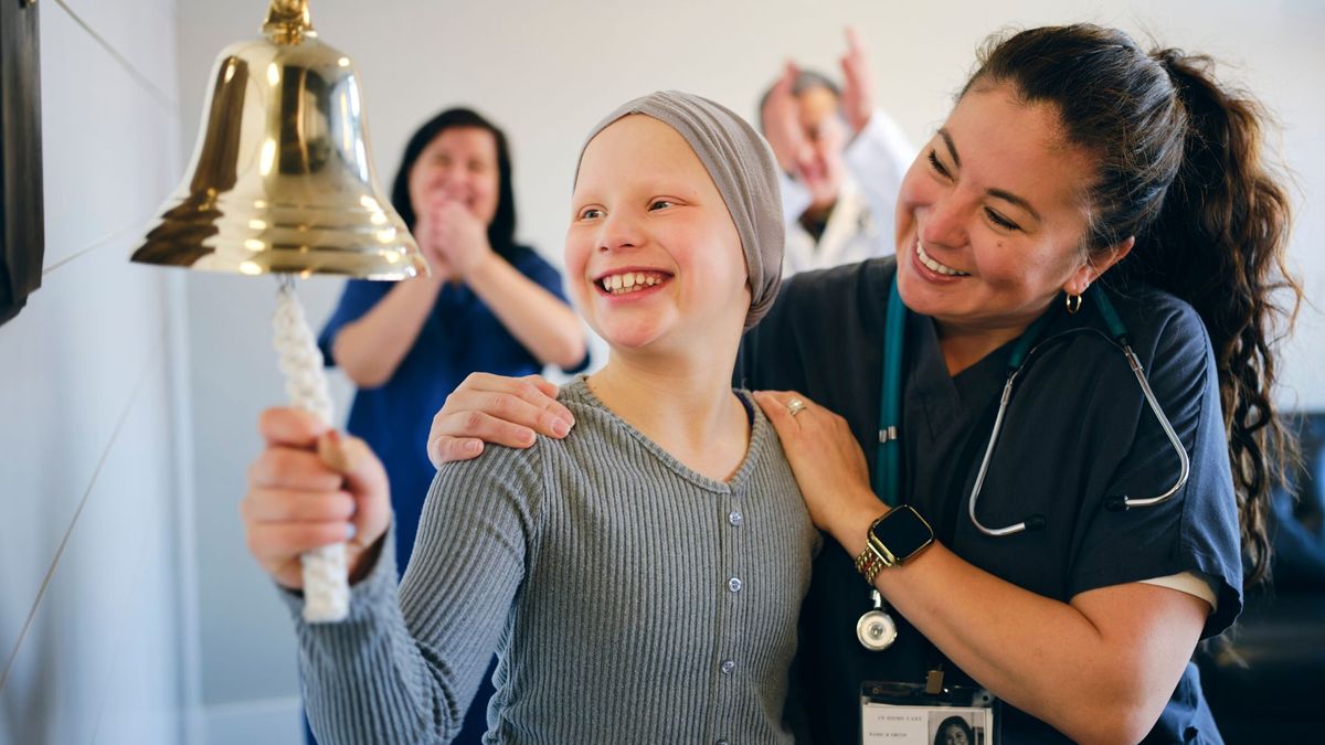 young girl with a scarf tied over her head rings a large bell while being hugged by a woman in a nurse&#039;s uniform; the scene suggests the girl just finished cancer treatment