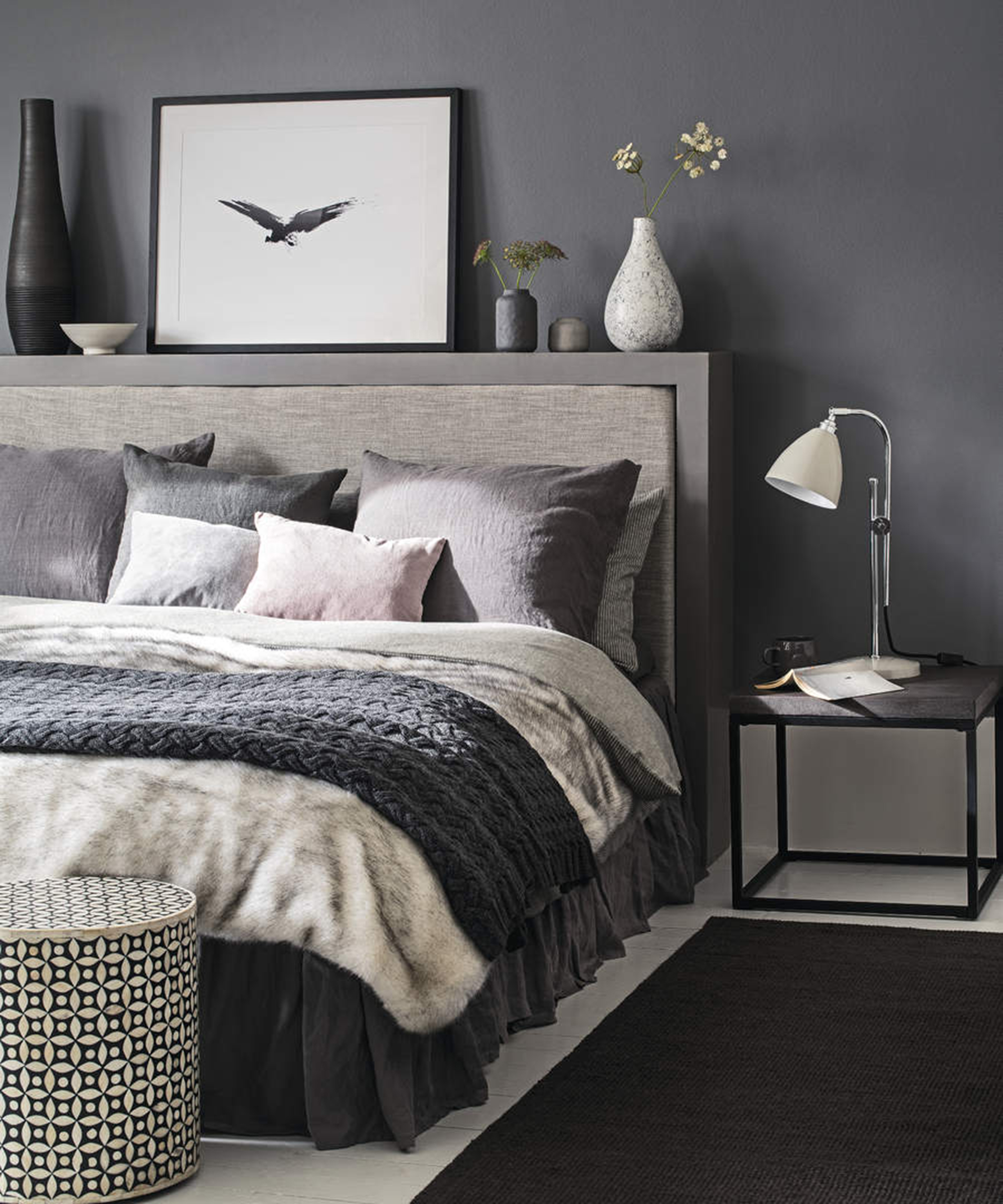 A grey bedroom with geometric patterned laundry basket, wood and metal side table and ornaments on raised shelf above headboard