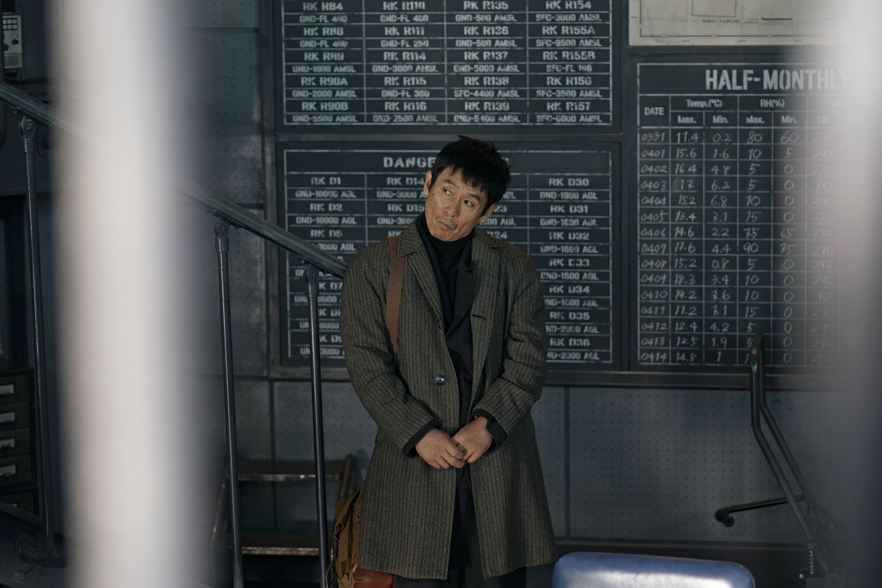 A man (Sul Kyung-gu as Mr. Nobody) stands in an abashed pose in front of blackboards filled with flight stats, in the Netflix Korean movie 'Good News.'