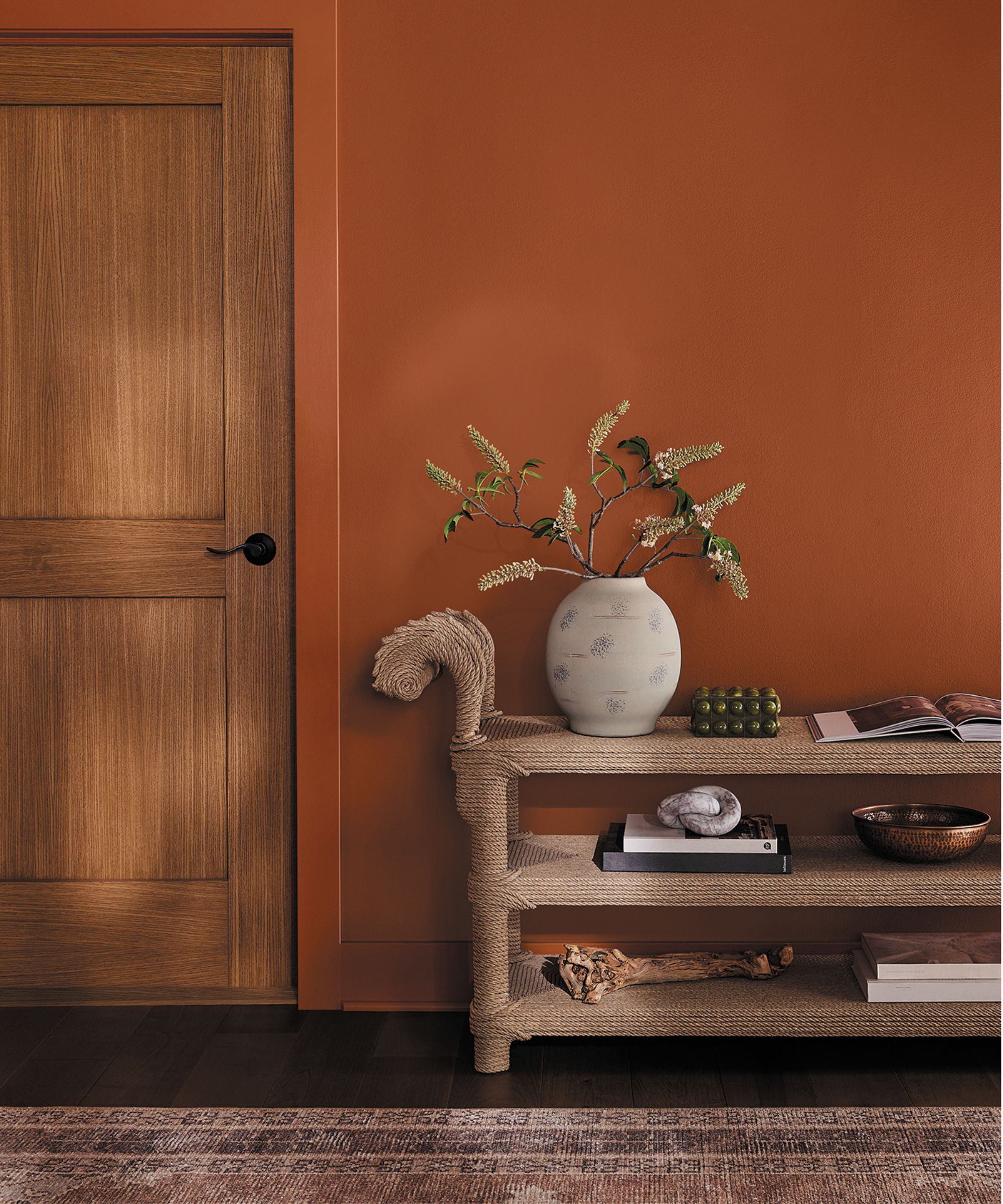 Hallway with orange walls, a wooden door, a rattan console holding stacks of books, trinkets, and a vase of flowers, with a dusty pink Persian rug below.