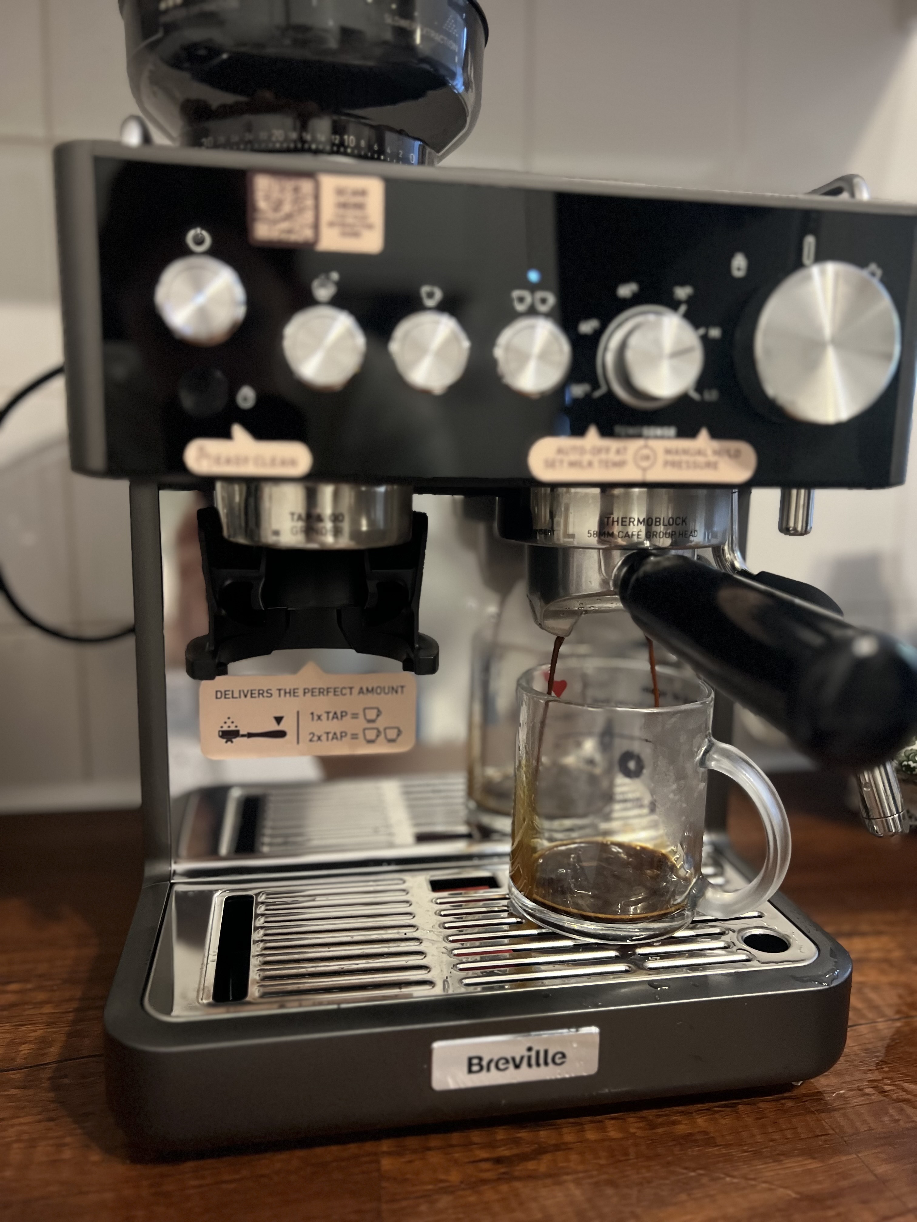 The Breville Barista Sense Espresso Machine in dark gray and black on a wooden countertop, brewing a shot of espresso in a glass mug.