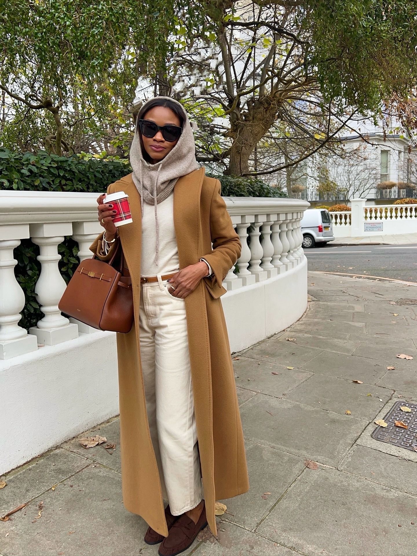 Lorna wears a beige balacava, white top, beige coat, white jeans, and brown loafers.