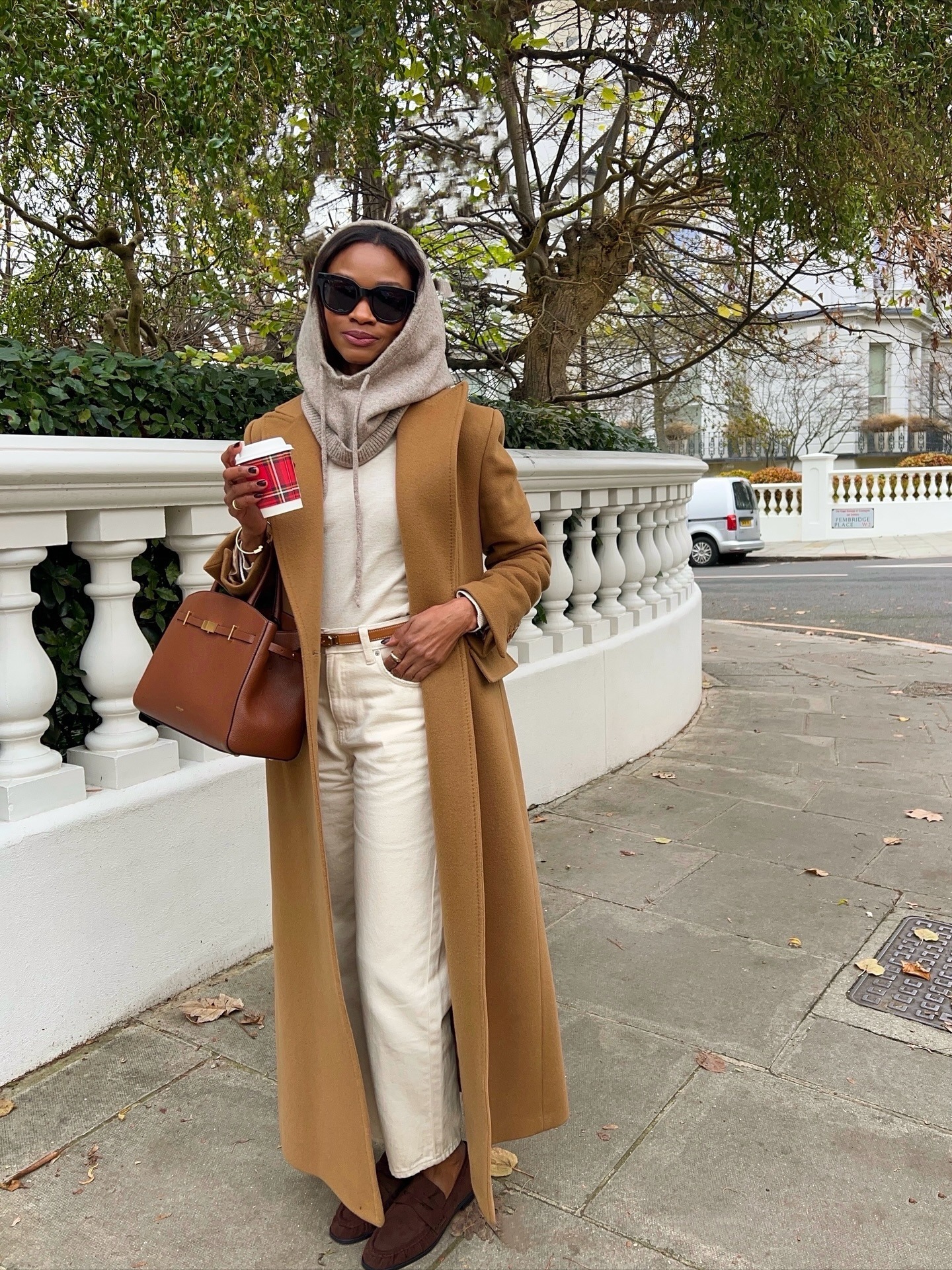 Lorna wears a beige balacava, white top, beige coat, white jeans, and brown loafers.