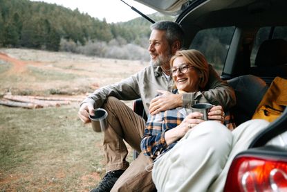 A mature couple takes a break after hiking by enjoying a hot drink by a car trunk in nature.