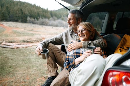 A mature couple takes a break after hiking by enjoying a hot drink by a car trunk in nature.