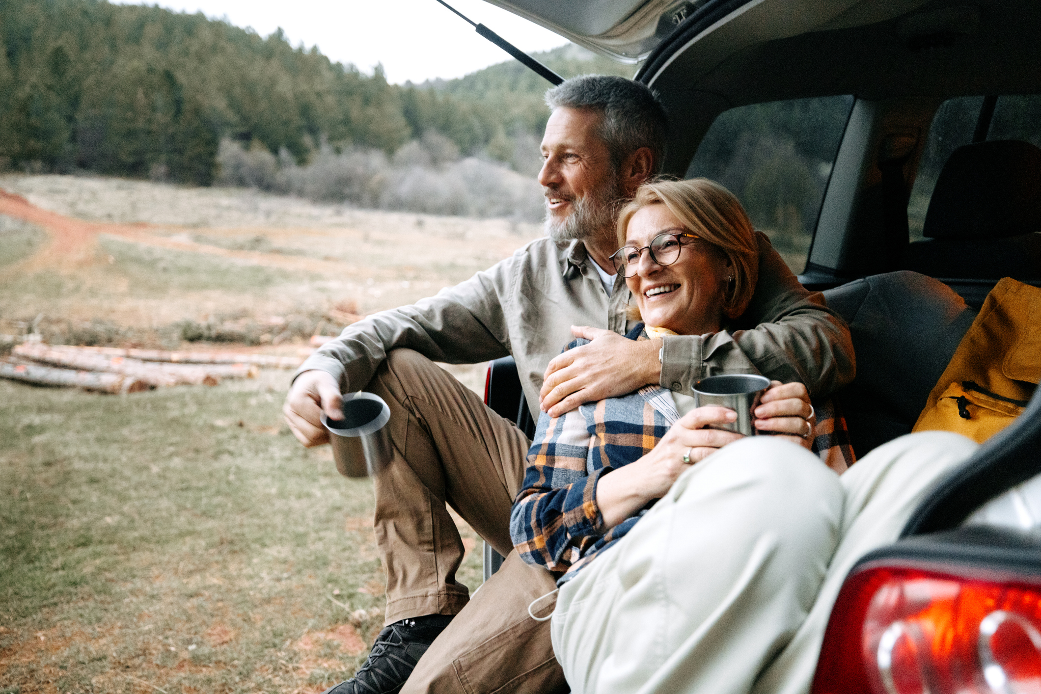 A mature couple takes a break after hiking by enjoying a hot drink by a car trunk in nature.