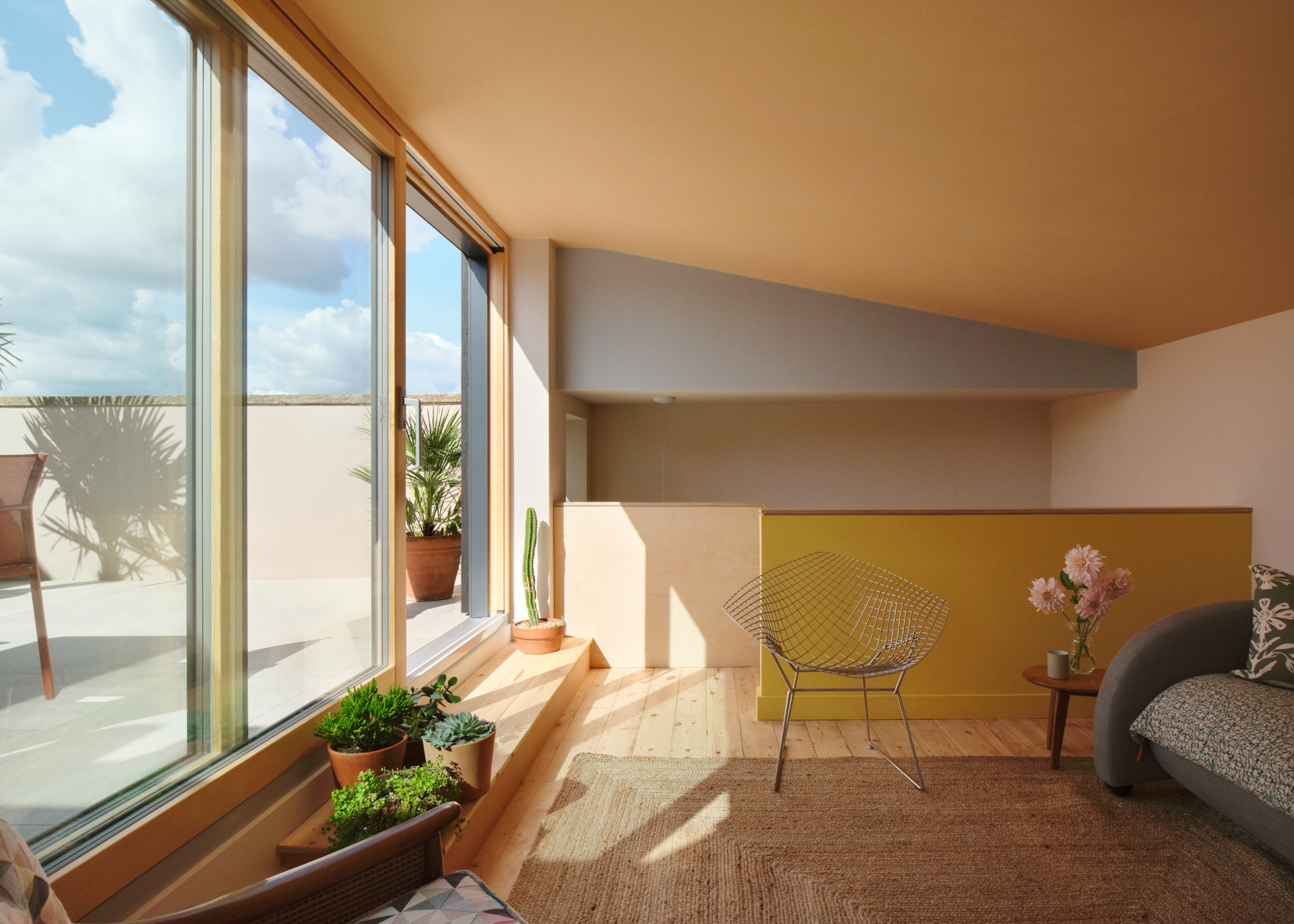 An extension space with sloped ceiling painted in warm joyful tones with sliding doors, a step to the outside space with plants on it and a mesh metal chair next to a small table with flowers