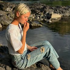 Iris Law sits by a lake wearing ripped jeans with a white boho blouse.