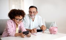 Money Skills. Smiling Senior Grandpa and Grandson Boy Engaging in Finance Education, Hold Wallet And Calculating Savings with Calculator Sitting Near Table With Piggybank At Home. Selective Focus