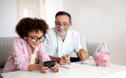 Money Skills. Smiling Senior Grandpa and Grandson Boy Engaging in Finance Education, Hold Wallet And Calculating Savings with Calculator Sitting Near Table With Piggybank At Home. Selective Focus