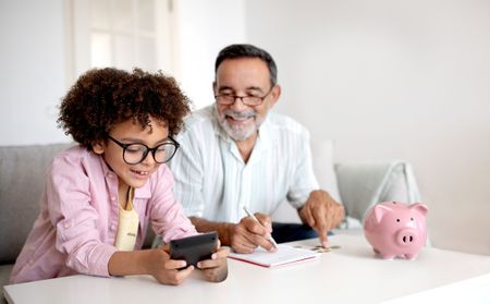 Money Skills. Smiling Senior Grandpa and Grandson Boy Engaging in Finance Education, Hold Wallet And Calculating Savings with Calculator Sitting Near Table With Piggybank At Home. Selective Focus