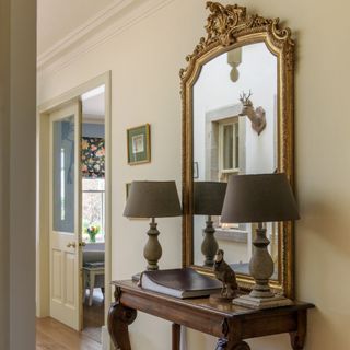 A hallway with a large gilded mirror and two large table lamps on a console table