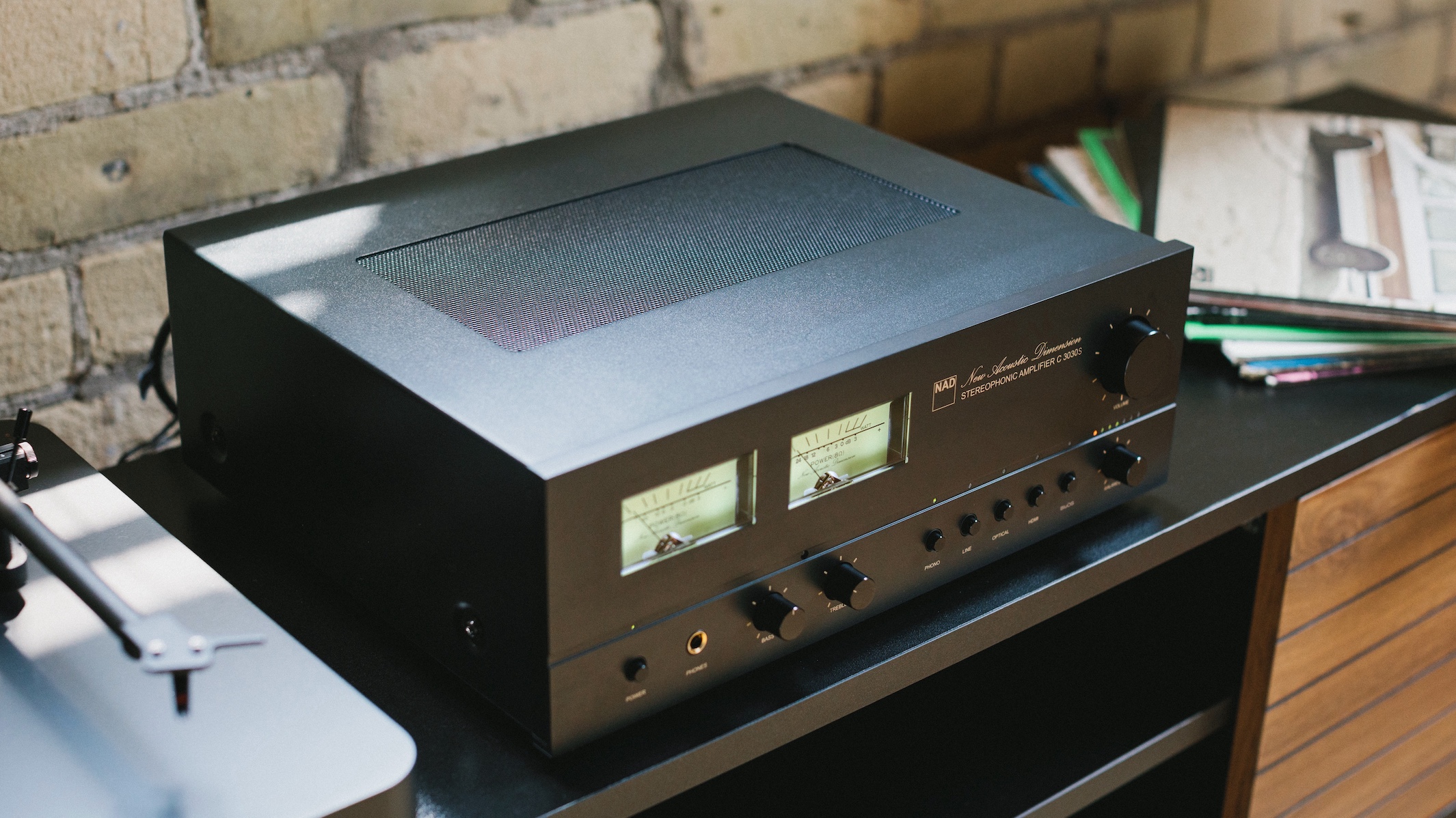 A NAD C 3030S integrated amplifier on a unit next to a record player in front of a brick wall, shot at a 3/4 angle.