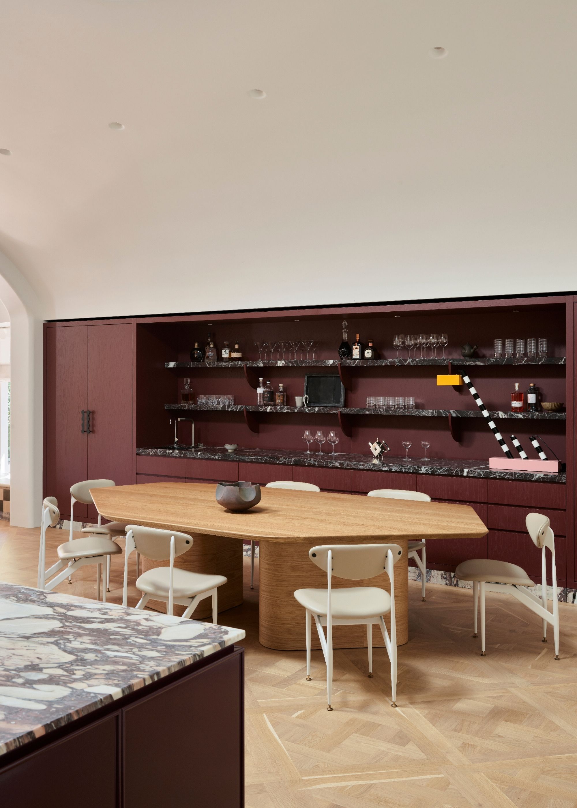 A modern kitchen with integrated storage in burgundy with marble detailing with open shelving and cupboards and drawers in front of a wooden dining table with cream chairs