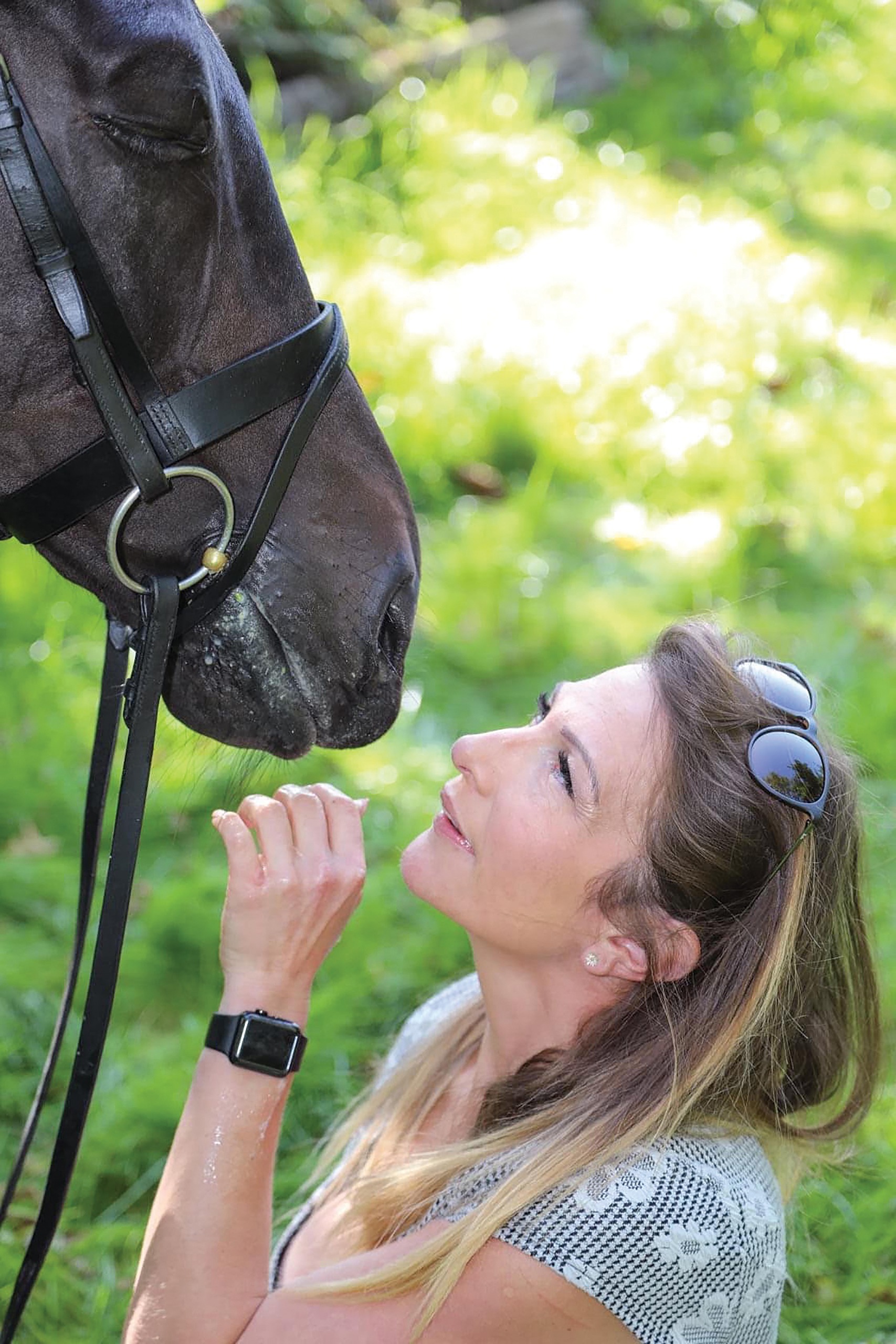 A lady who is kneeling looks up at a dark bay horse's head. The horse is wearing a flash bridle and has his eye closed.