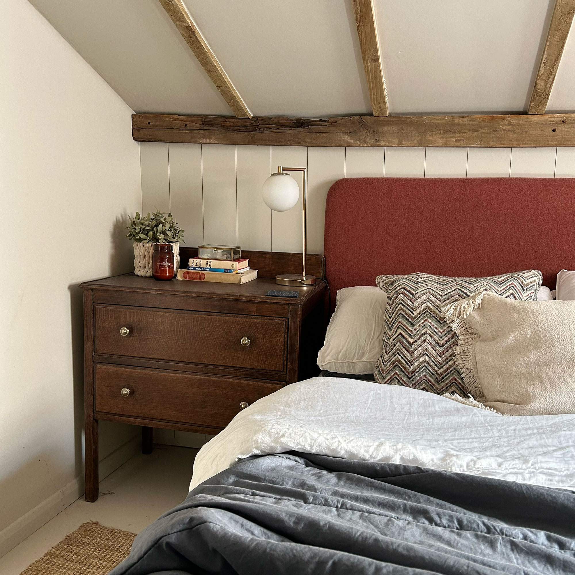 Bed with red fabric headboard beside wooden bedside table underneath eaves ceiling with wooden ceiling beams