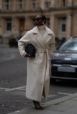 A cute work winter outfit is shown in a photo of a woman standing on the sidewalk in London wearing a long white maxi coat over a white sweater dress with gold earrings, black sunglasses, a black Prada bag and black slingback heels