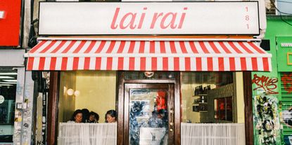 A small restaurant's exterior features a red and white tent, a sign reading "lai rai" and "181" red on white, bistro-style curtains, and buttery interiors with spherical sconces and people seen chatting.
