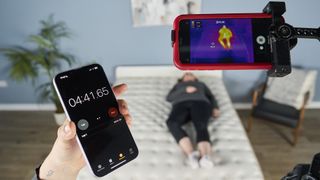An image showing behind the scenes of our temperature regulation tests on the Saatva Classic mattress, with our tester lying on the mattress and our Managing Editor of Sleep, Claire Davies' iPhone attached to our thermal imaging camera to track the heat absorption and heat dissipation capabilities of the Saatva Classic over a period of 30 minutes.