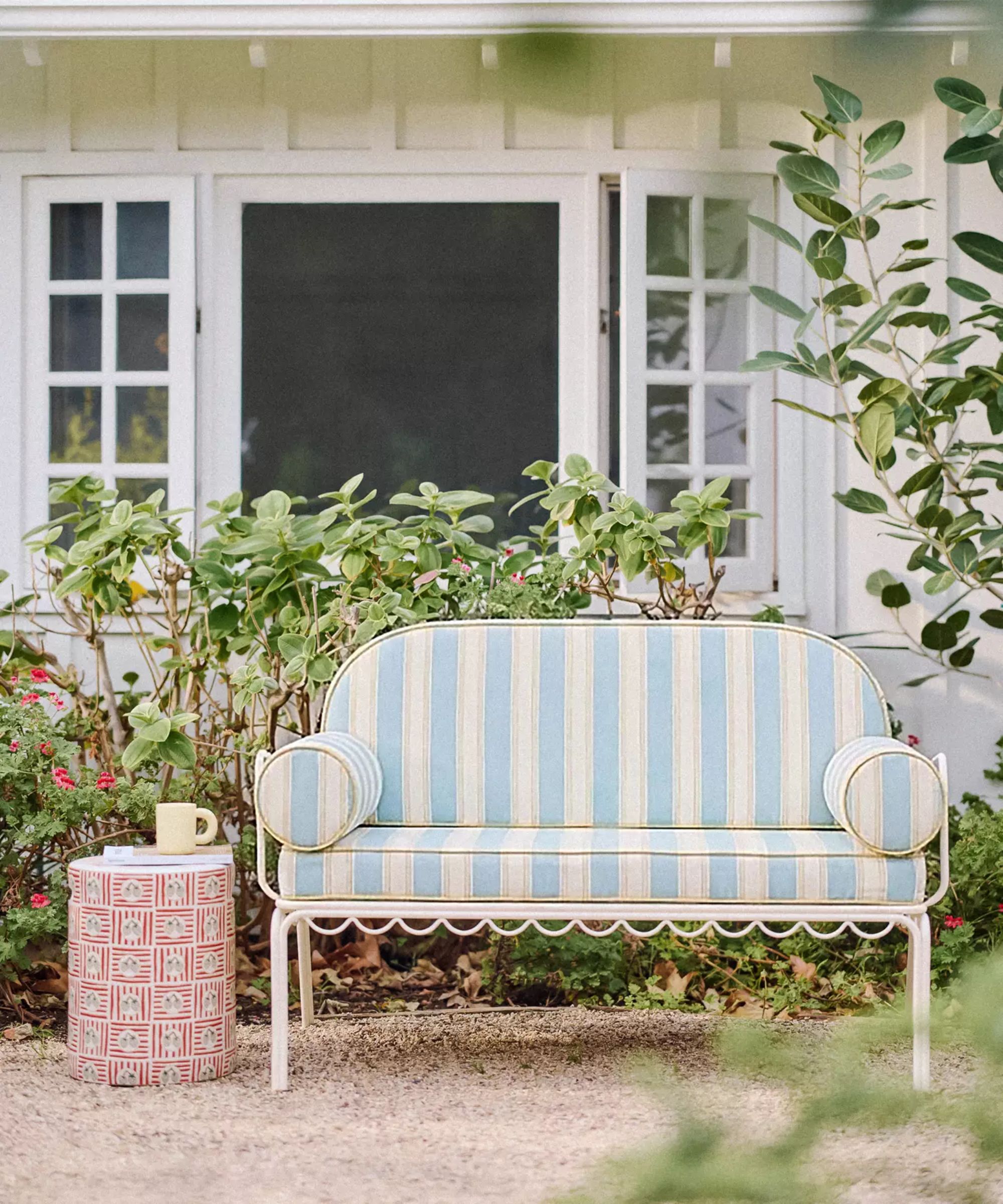 a garden with a patio clad garden room and a small white wrought iron sofa with scalloped detail and striped blue cushions