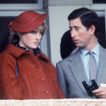 Princess Diana wears a red mohair coat with a matching hat while King Charles wears a light gray suit with a blue shirt and looks at his wife with a worried expression